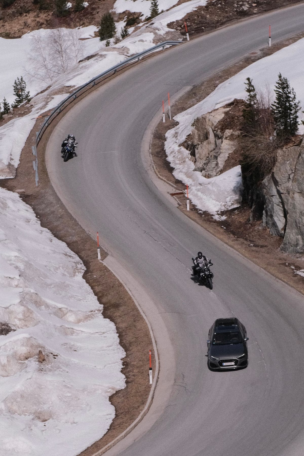 Dve motorky a auto v zákrute horskej cesty so snehom na krajoch
