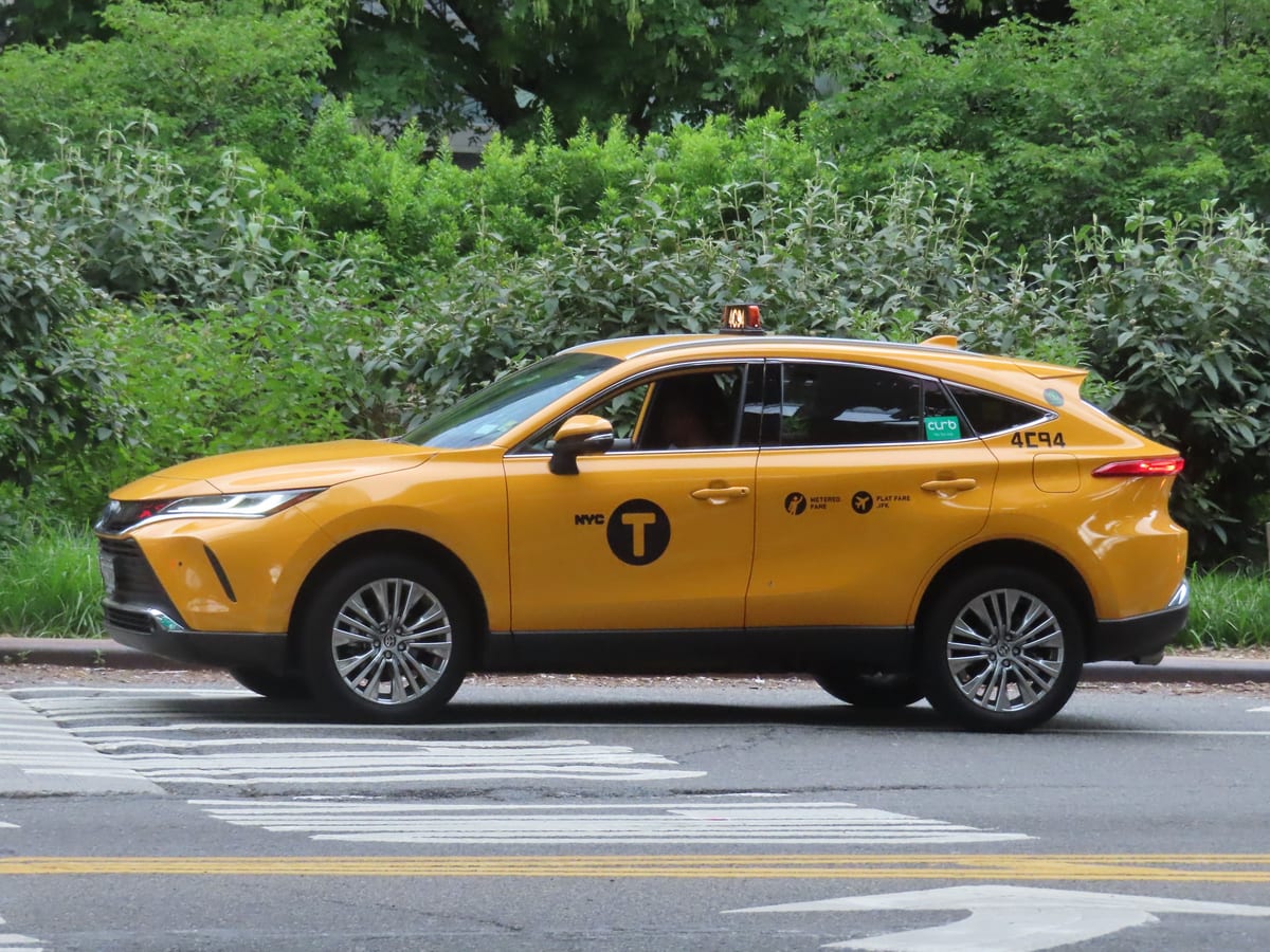 Žltý newyorský taxík Toyota Venza so znakom NYC Taxi na ulici so zeleňou v pozadí