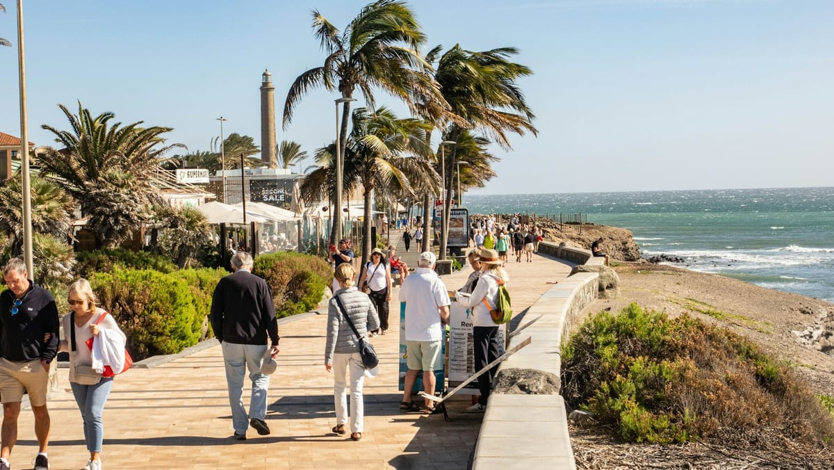 Ľudia na promenáde pri majáku Maspalomas, palmy sa ohýbajú vo vetre a vpravo je more.