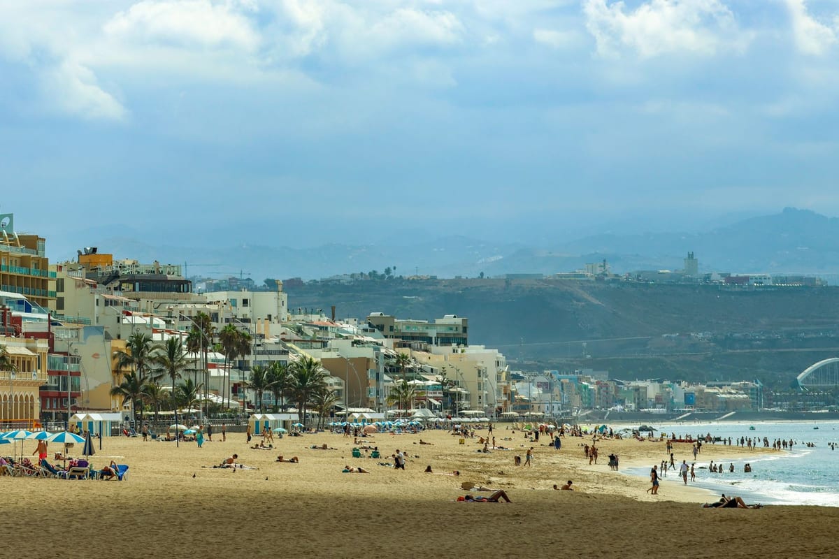 Slnečná pláž Las Canteras v Las Palmas s ľuďmi na piesku a domami pozdĺž promenády.