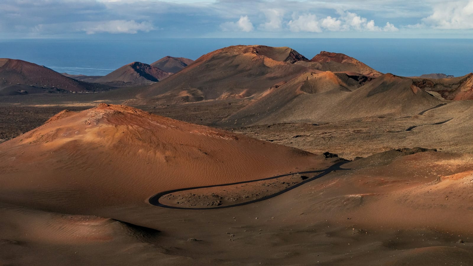 Červenohnedé sopky národného parku Timanfaya s cestou, ktorá sa kľukatí krajinou, a oceánom v pozadí