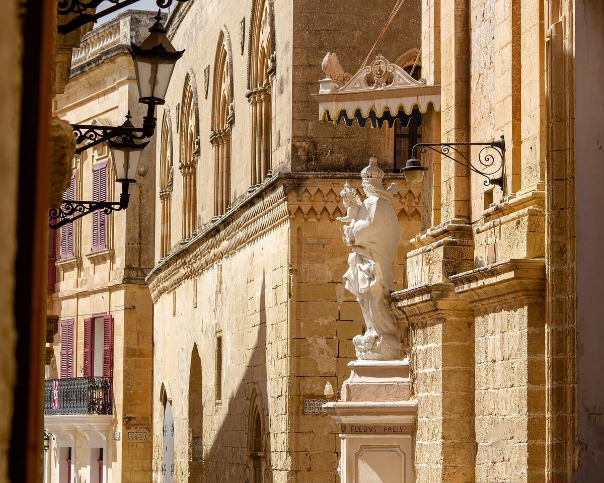 Kamenné paláce so sochou a lampami v úzkej ulici opevneného mesta Mdina
