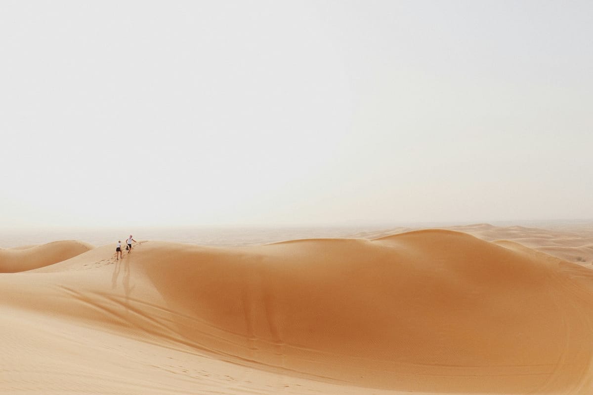 Dvaja ľudia kráčajú po vysokej piesočnej dune v púšti pri Dubaji pod bledou oblohou