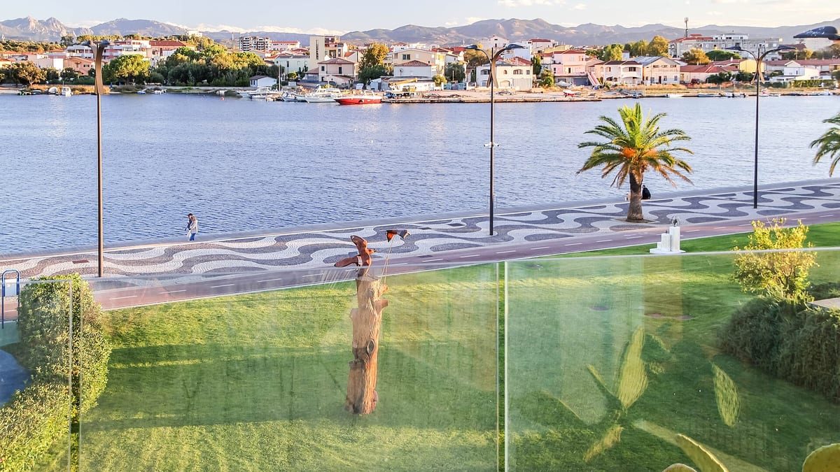 Promenáda Lungomare v Olbii s palmou a vodou zálivu, na druhom brehu domy