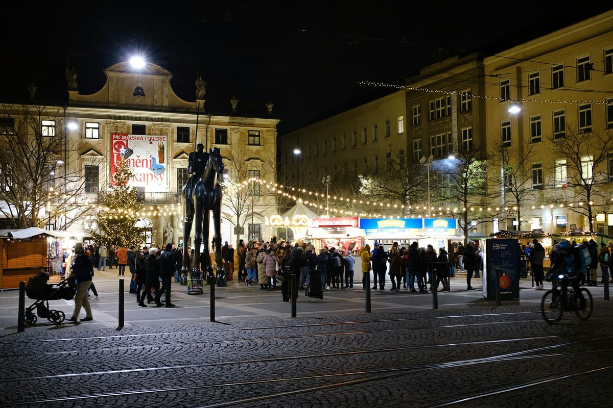 Večerný vianočný trh pri soche Jošta na Moravskom námestí v Brne so svetielkami nad stánkami