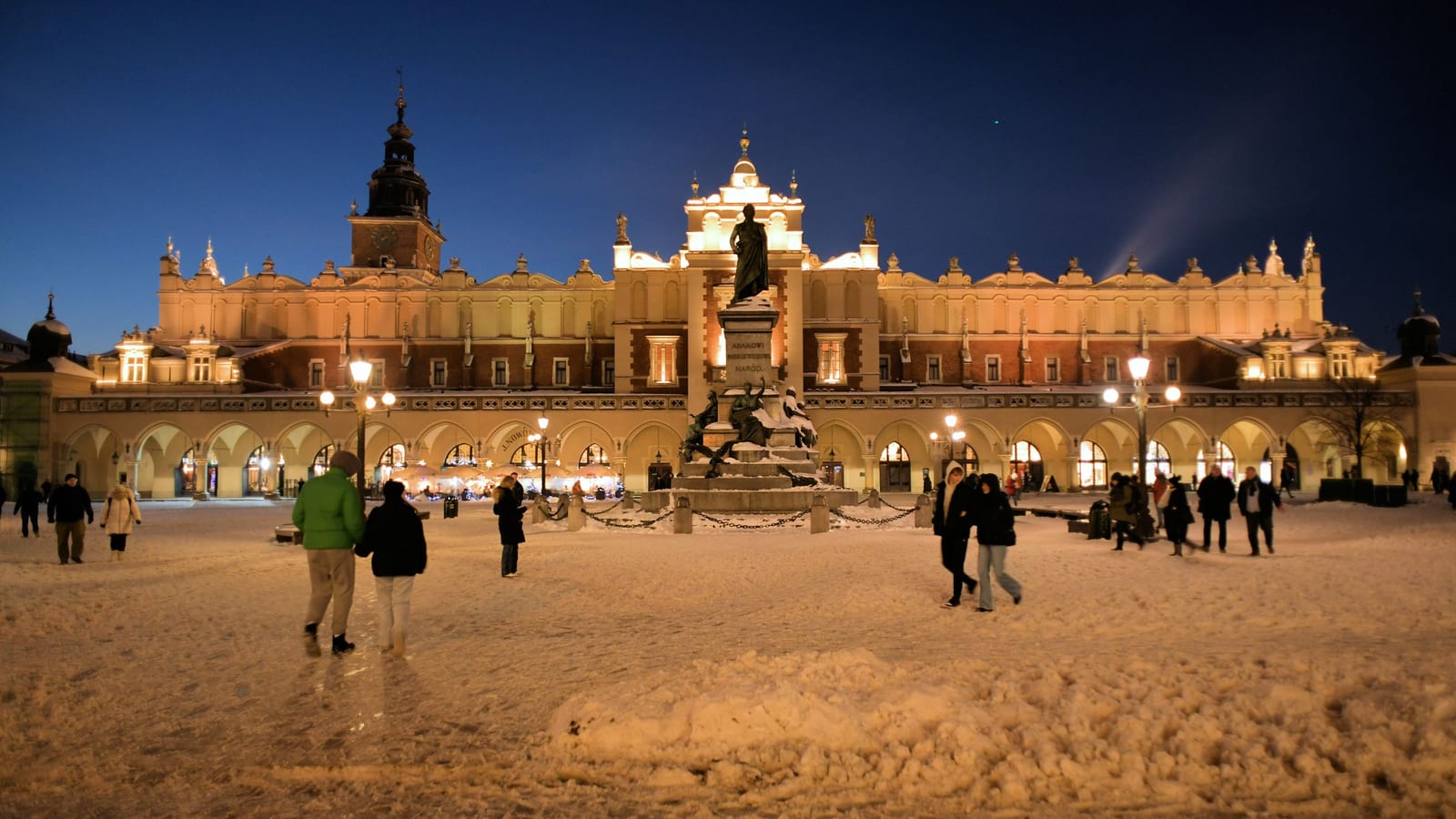 Zasnežený Rynek Główny v Krakove večer s osvetlenými Sukiennicami a ľuďmi na námestí