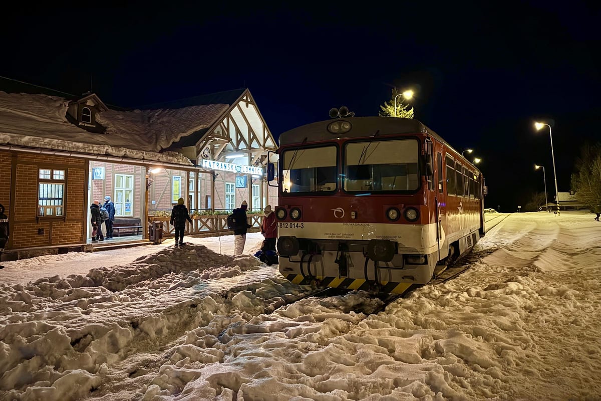 Drevená staničná budova v Tatranskej Lomnici a motorový vlak večer medzi kopami snehu.
