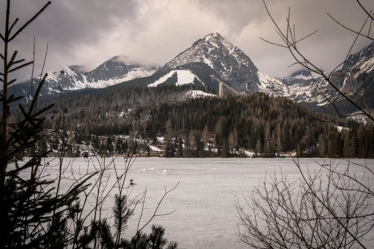 Zamrznuté Štrbské pleso v zime, za lesom skokanské mostíky a zasnežené štíty Vysokých Tatier.