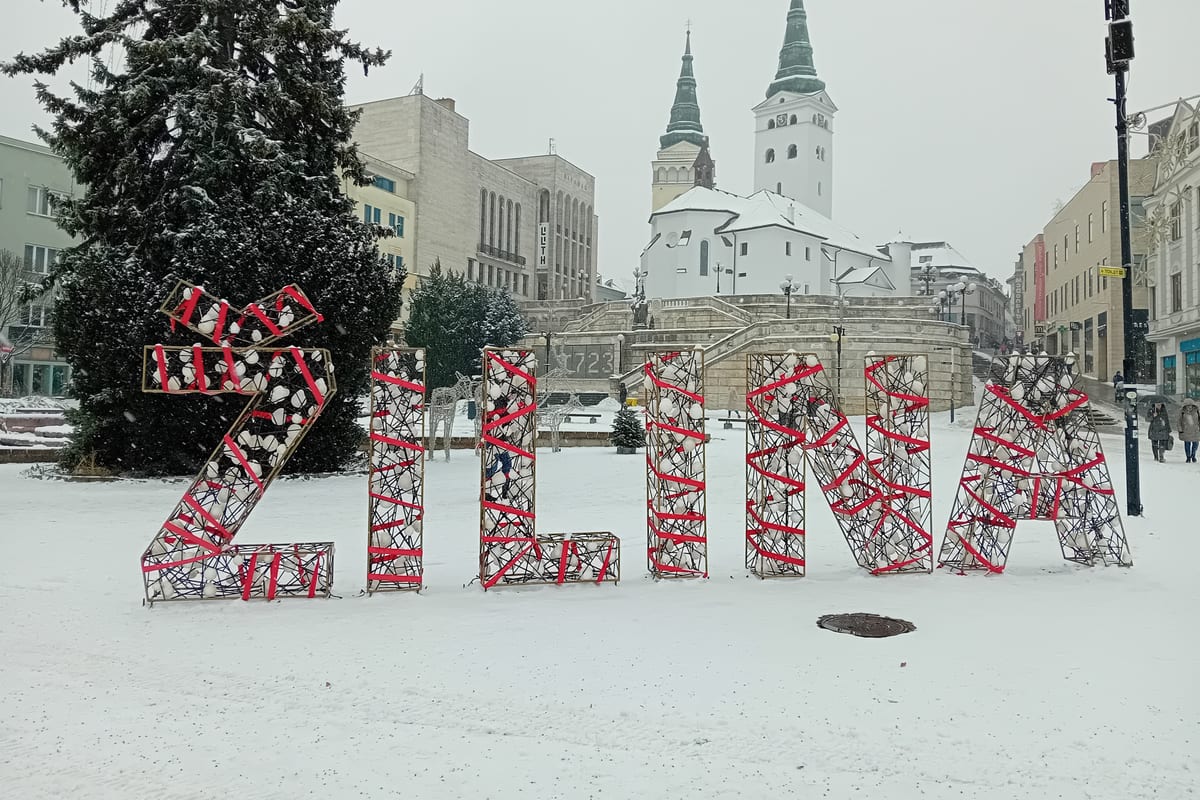 Veľký nápis ŽILINA a vianočný stromček v snehu, v pozadí Katedrála Najsvätejšej Trojice a Burianova veža.