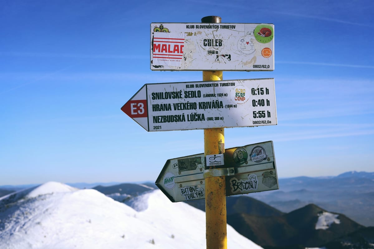 Turistický rozcestník na vrchole Chleb v Malej Fatre so zasneženým hrebeňom a smerom k lanovke na Snilovskom sedle.